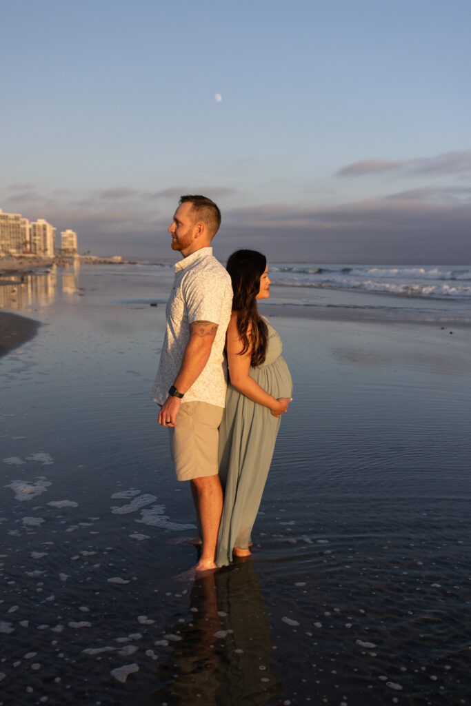 Couple stands back to back at coronado beach, with the wife holding her pregnant stomach